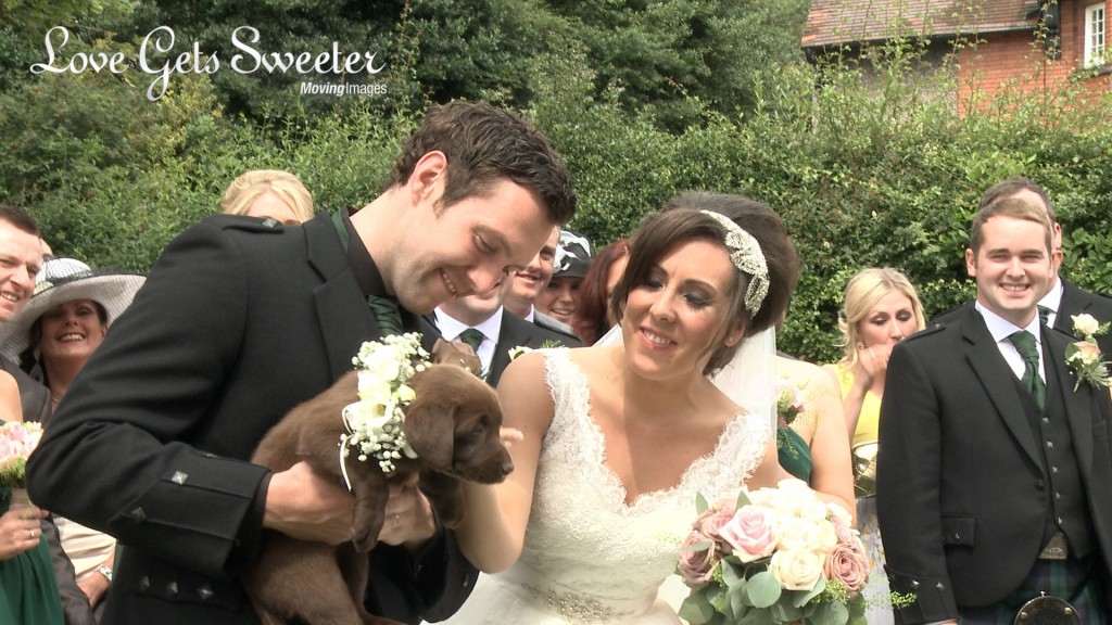 Vicky and Gareth Highlights20 copy The Groom shows off his surprise to the bride for the wedding videographer and it's a chocolate Labrador wearing a cream rose flower collar outside their church in the Wirral