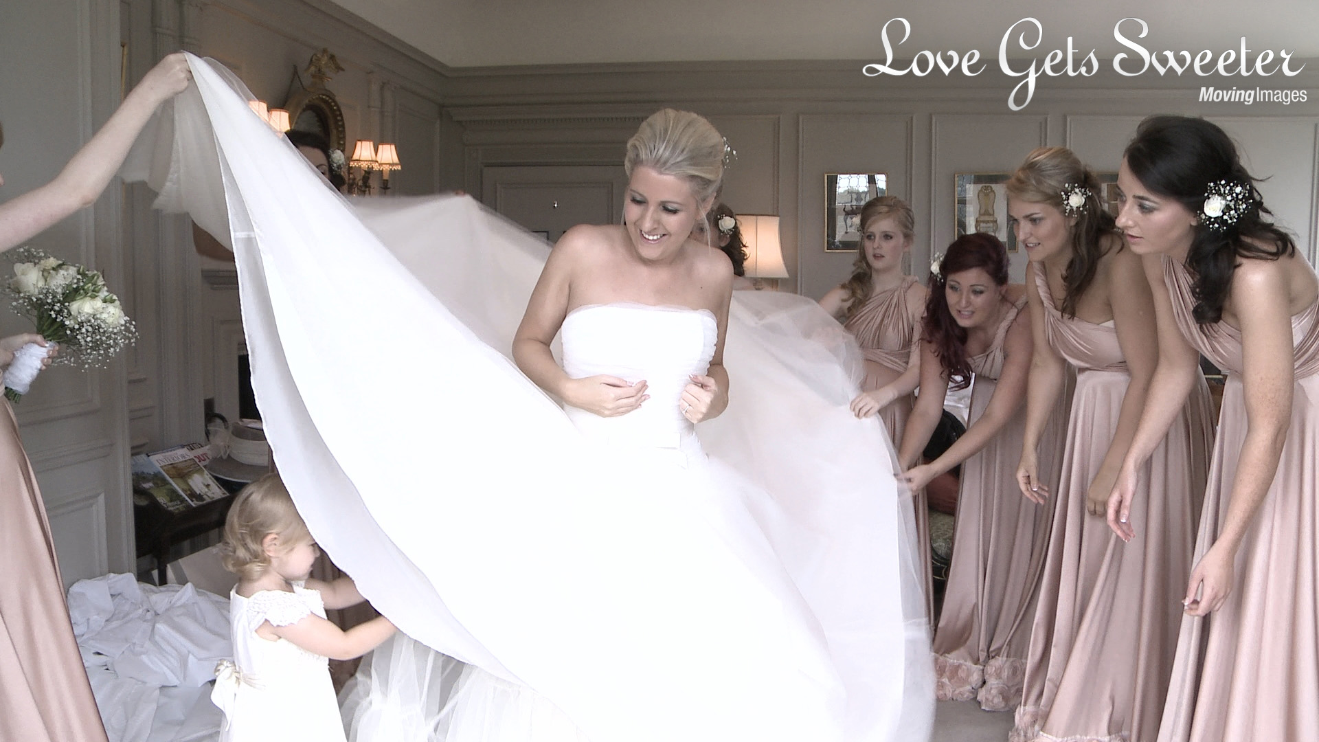 bride in Stewart Parvin wedding gown with her bridesmaids in two bird pink bridesmaid dresses watching the cute flower girl help under the brides dress at thornton manor in the wirral