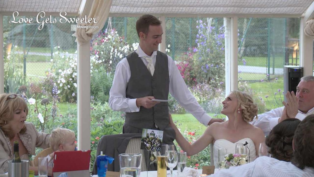 The groom has his hand on his brides shoulder during his wedding speech on the wedding video. They smile at each other as they do speeches at the top table at their Abbeywood Estate wedding