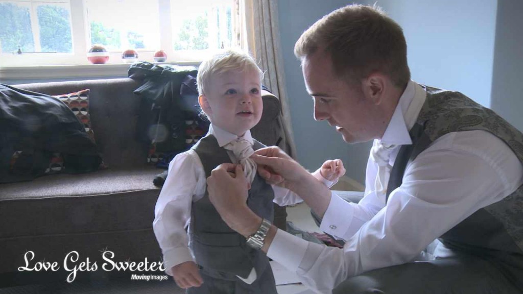 the groom is also a dad and he kneels down to fix his toddler sons wedding tie as they get ready with the wedding videographer at Abbeywood Estate and Gardens in Cheshire