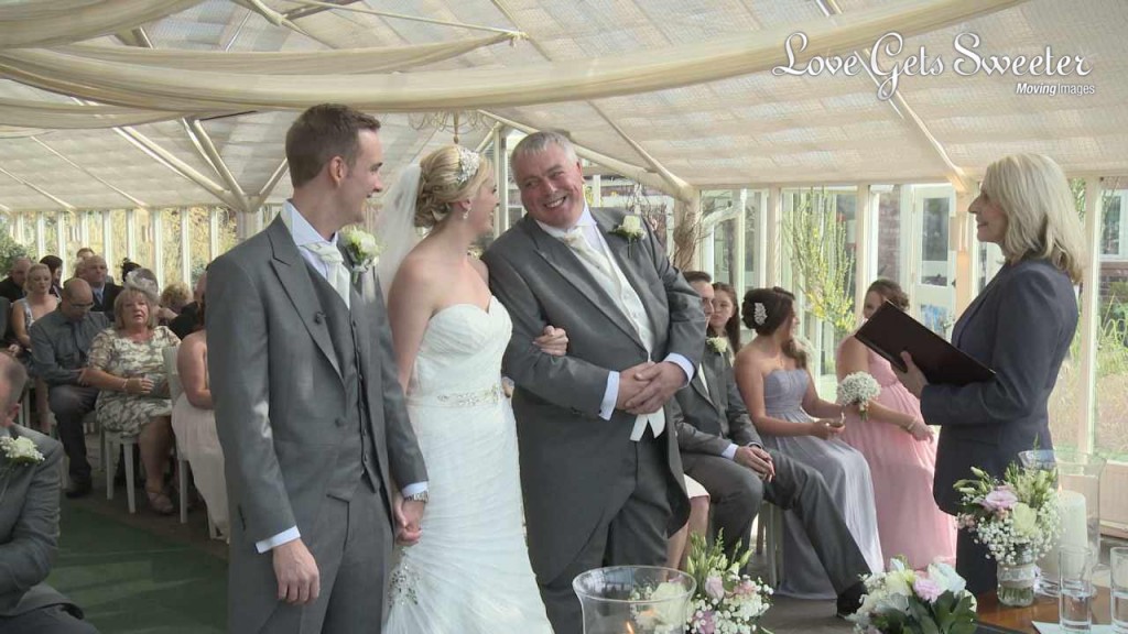 A wedding video still of a father of the bride leans over and smiles proudly at his daughter who has linked her arm in his after walking her down the aisle at Abbeywood Estate in Cheshire