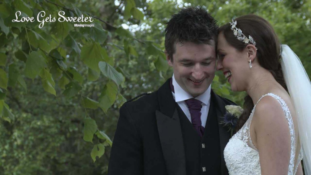 A video still of the bride leaning against the groom in his Scottish wedding outfit in front of the green leaves at Mere Court Hotel. Showing off her hair piece and big beautiful smile