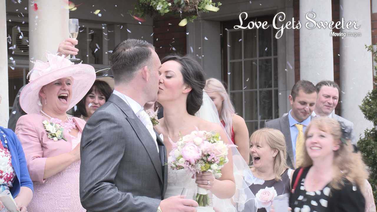 A bride and groom kiss outside the front of the historic looking manor house that is Eaves Hall in Clitheroe and kiss as their guests throw confetti for the photographer and videographer