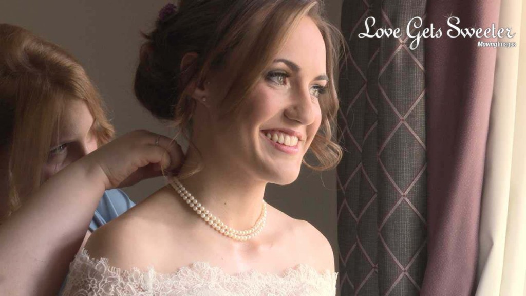 Charlotte the bride gives a huge smile on the wedding video as her maid of honour helps add her pearls to finish off her wedding day look at Rookery Hall