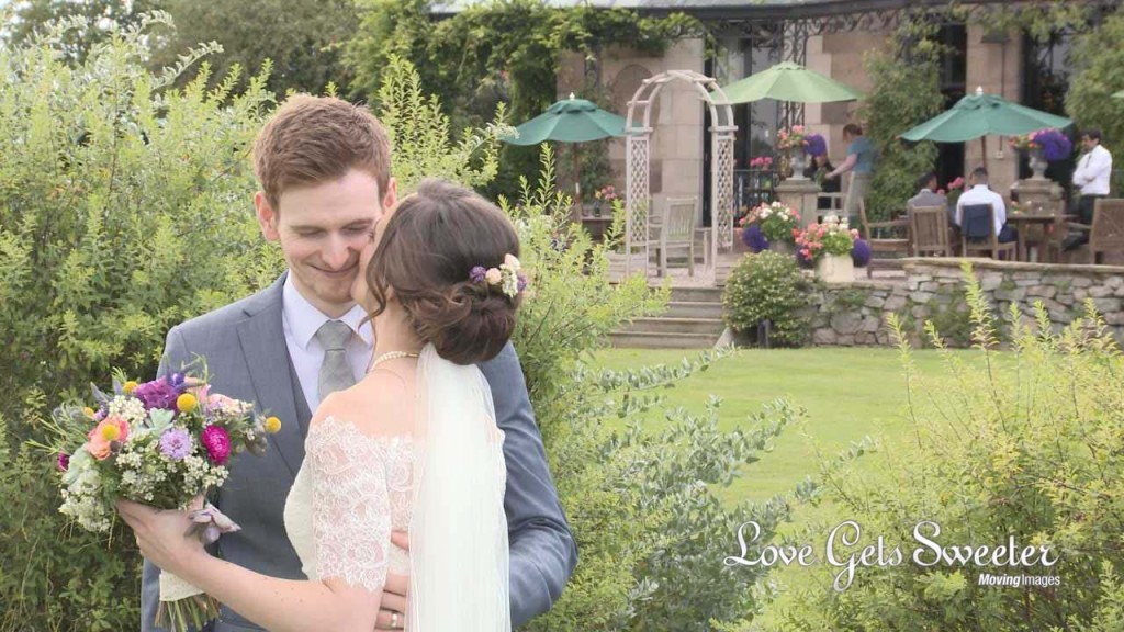 the groom smiles as his bride leans in for a kiss outside their wedding venue rookery hall. She holds a vintage inspired colourful bouquet of pinks, oranges, yellow and purple