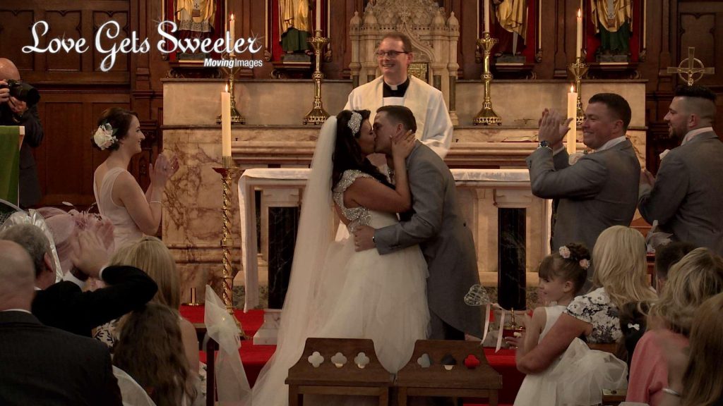 A wide shot from the wedding video as the bride and groom kiss and all the guests clap and cheer for the end of the wedding service at St Peters Church in Cheshire