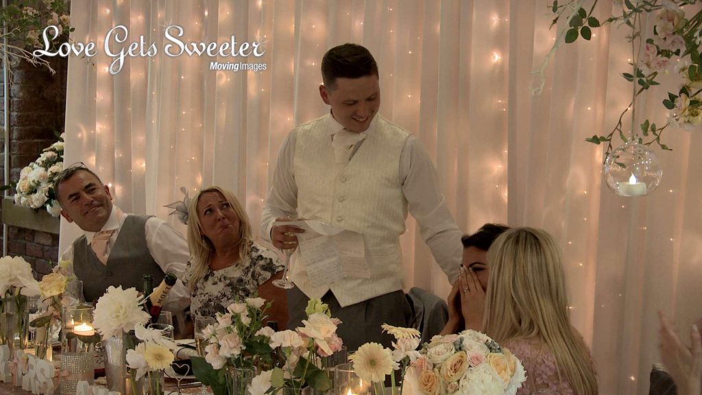 a wedding video still of the Bride getting emotional during the grooms romantic wedding speech and his parents looking on very proud. Their table has an informal and pretty cream and peach flower display as well as a sparkling fairy light back drop for their cheshire wedding