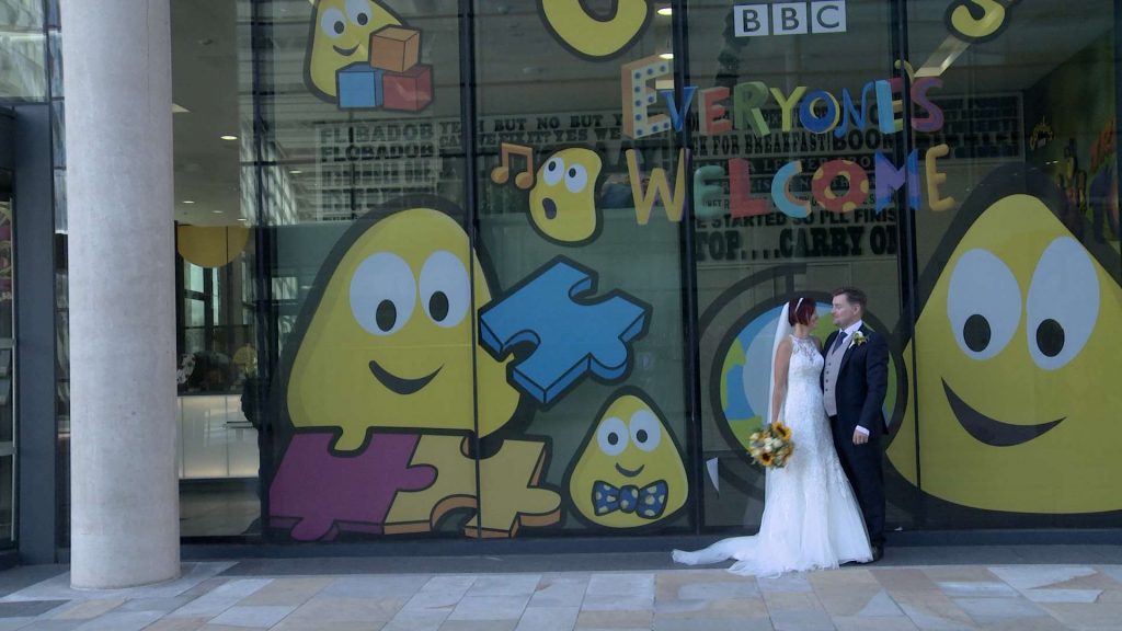 A bride holding a bright yellow sunflower bouquet smiles at her groom in front of the fun and colourful CBeebies window in Salford Media city