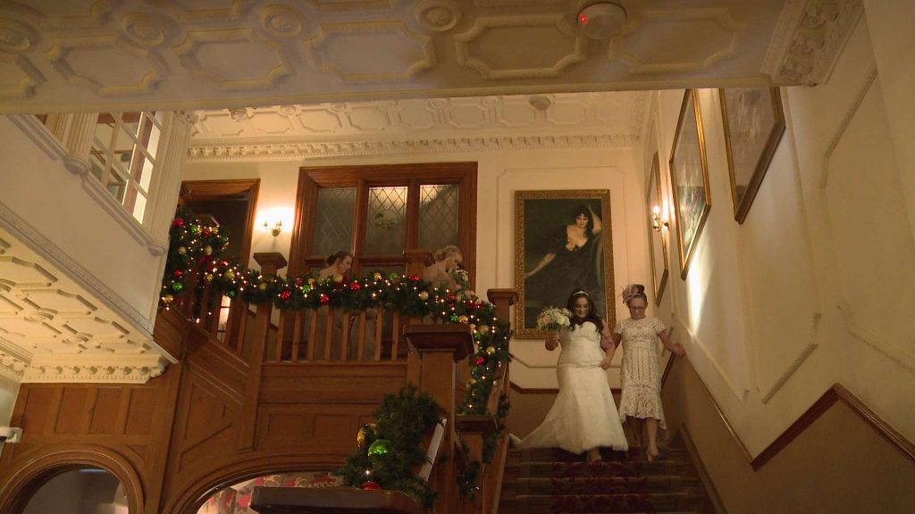 The bride walks down the grand oak staircase with her Mum. It's decorated with christmas garlands at Nunsmere Hall
