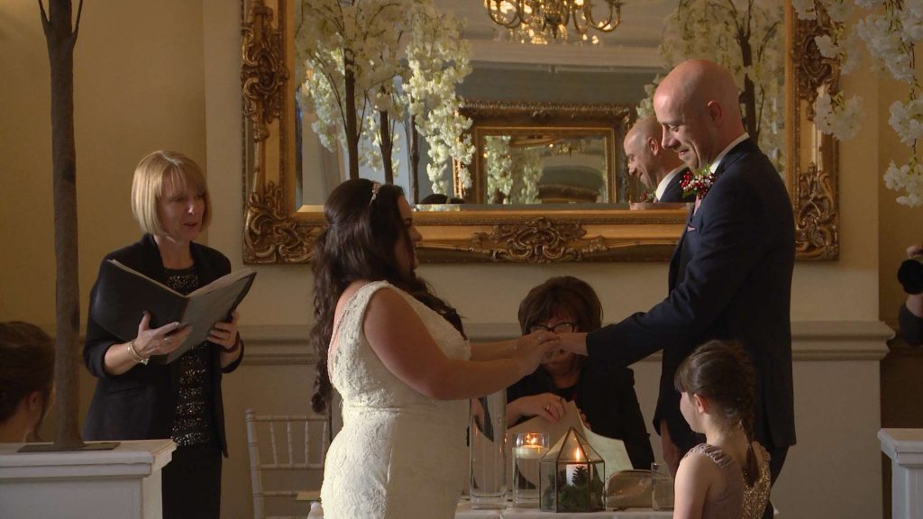 The bride laughs as she squeezes the wedding ring on to her groom on their wedding video