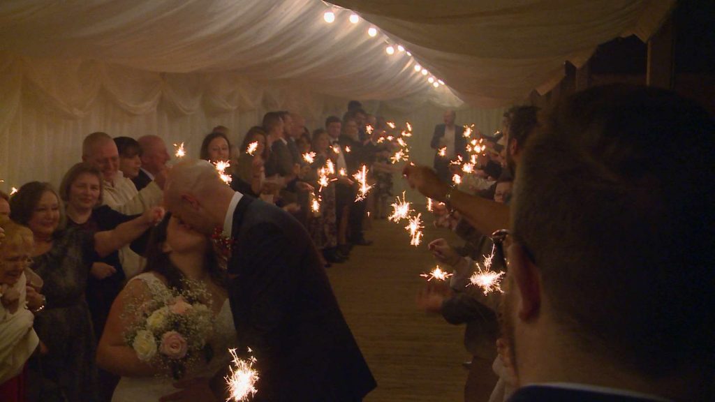 a bride and groom kiss as guests wave sparklers around and cheer for the video at Nunsmere hall