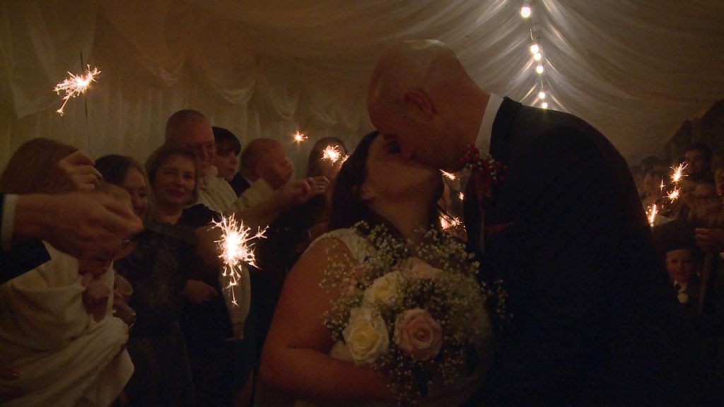 the bride and groom kiss in the marquee walkway surrounded by guests and sparklers at Nunsmere hall in Cheshire