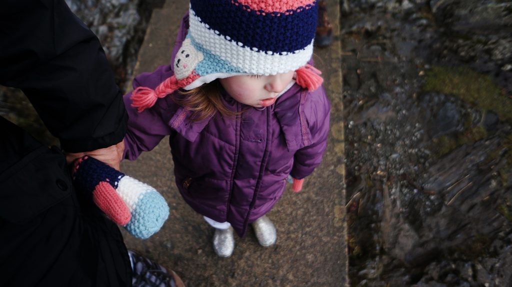 3 year old girl stands waiting for a boat at derwent water in her warm purple coat and matching polar bear hat and gloves
