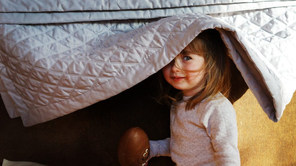 a little toddler girl smiles as she tried to hide herself and her easter egg under a silver throw hanging over a sofa as her Mum takes a photo in their holiday house in Penrith