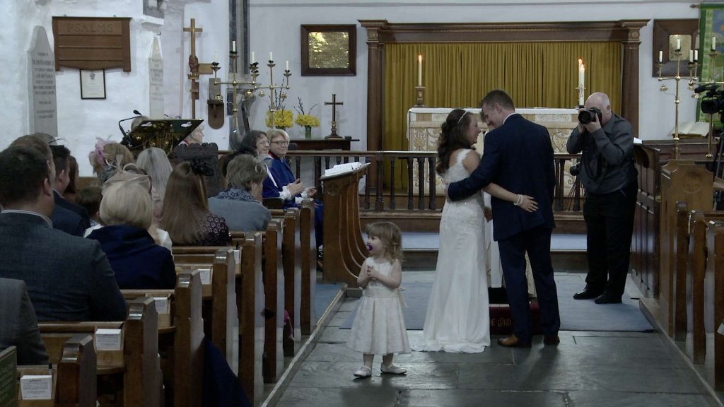 a flower girl stands in the middle of the aisle watching everyone clapping for her Mum and Dad getting married