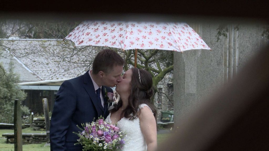a bride and groom enjoy a kiss under a white and pink flowery umbrella. the video camera is looking through the old wooden church gate at St Oswalds church in the lake district