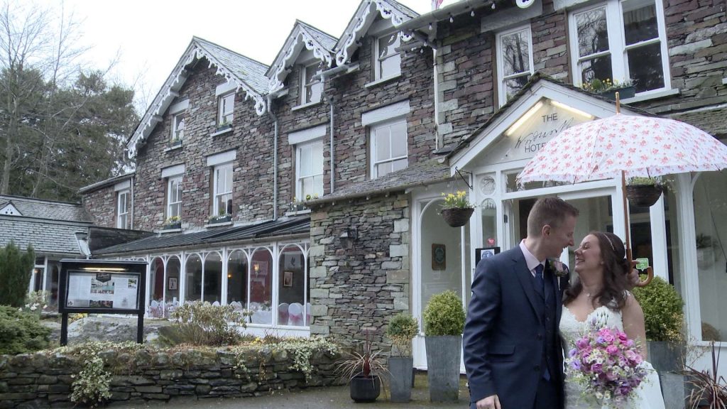 a bride and groom pose fro the wedding video under a pink and white flowery umbrella in front of the grey stone building that is The Wordsworth Hotel in Grasemere