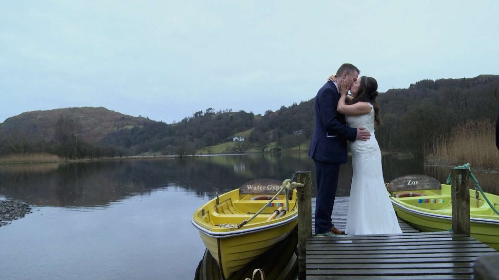 a bride and groom stand on a jetty with two bright coloured rowing boats by the side of Grasmere lake after their wedding in the lake district having a little kiss in the rain for their wedding photos and video