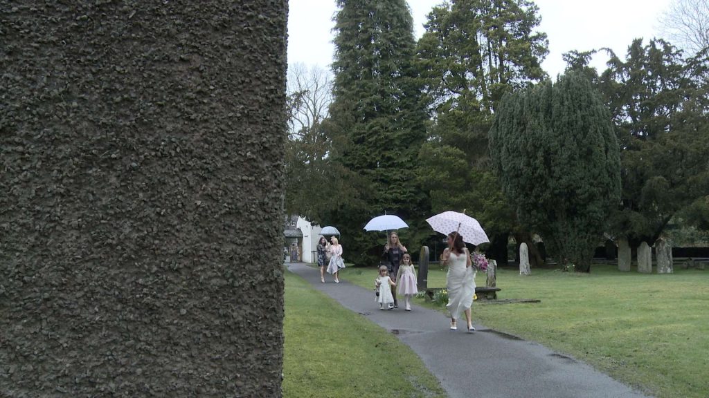 a wedding video still of a bride and her flower girl hold umbrellas as they walk in the rain through the gardens at St Oswalds in Grasmere before her wedding ceremony