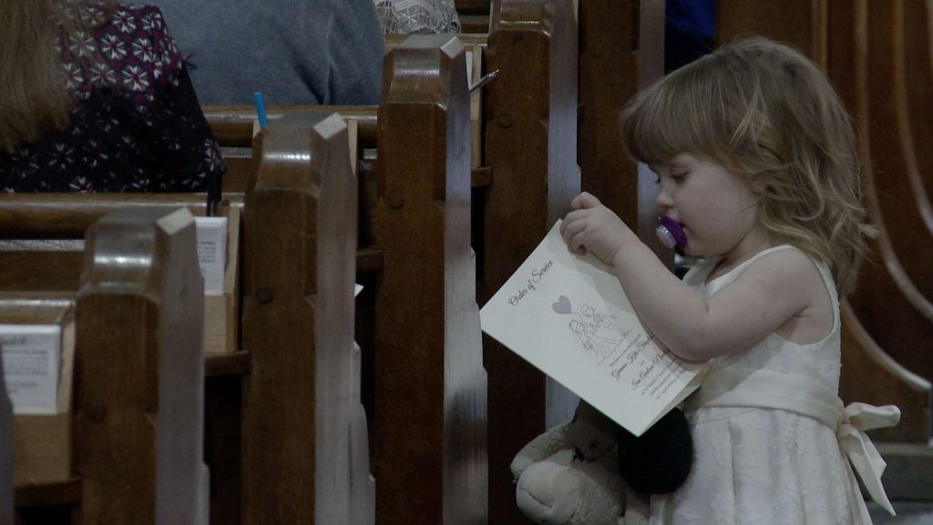 a little flower girl with a dummy in her mouth curiously opens up the order of service for her mummy and daddys wedding at St Oswalds church in Grasmere