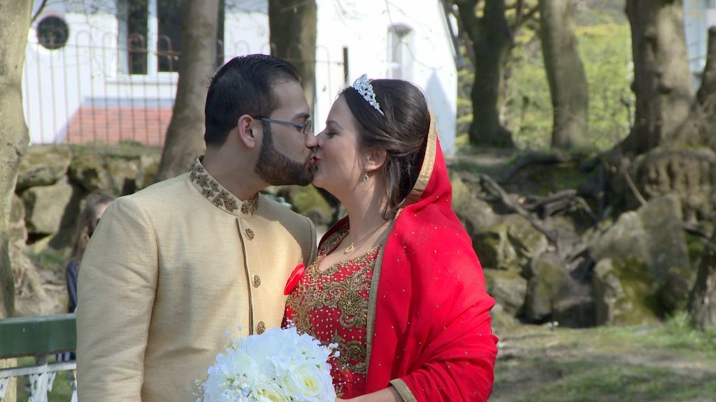 the bride and groom share a kiss in the dappled sunlight at southports botanic gardens. she wears a bangladeshi wedding saree in bright red and holds a simple white bridal bouquet