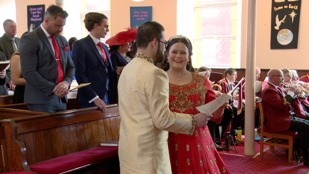a bride and groom smile at each other as they sing hymns inside church caught on their wedding video in Southport