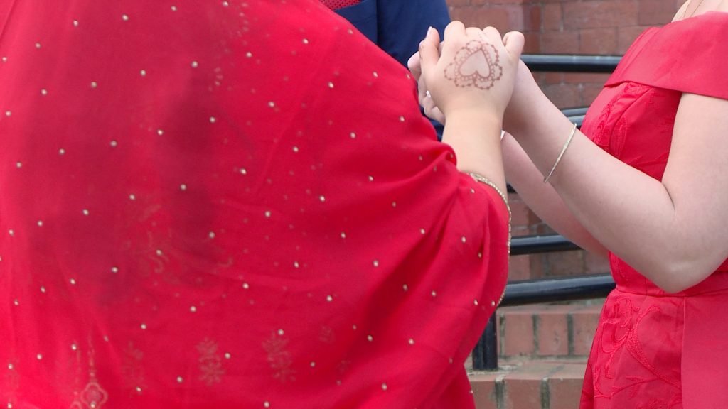 a close up of the brides bangladeshi dress with gold details and her heart shaped brown henna on her hand as she holds her sister and bridesmaids hands outside church