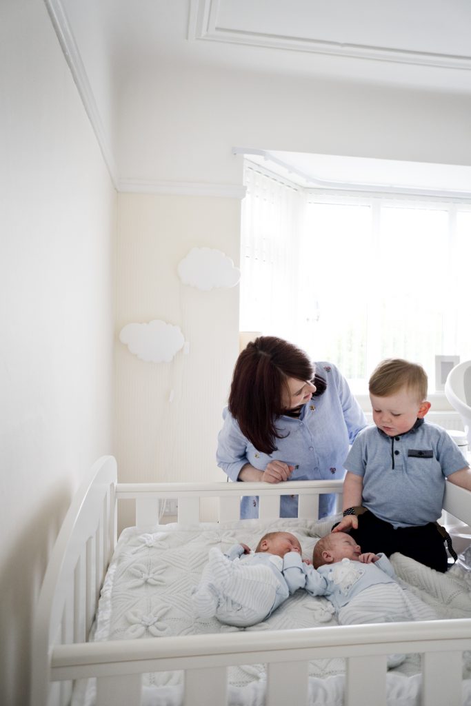 a mum leans over to chat to her toddler about their new baby boy twins in their bright and neutral white and cream nursery in the wirral