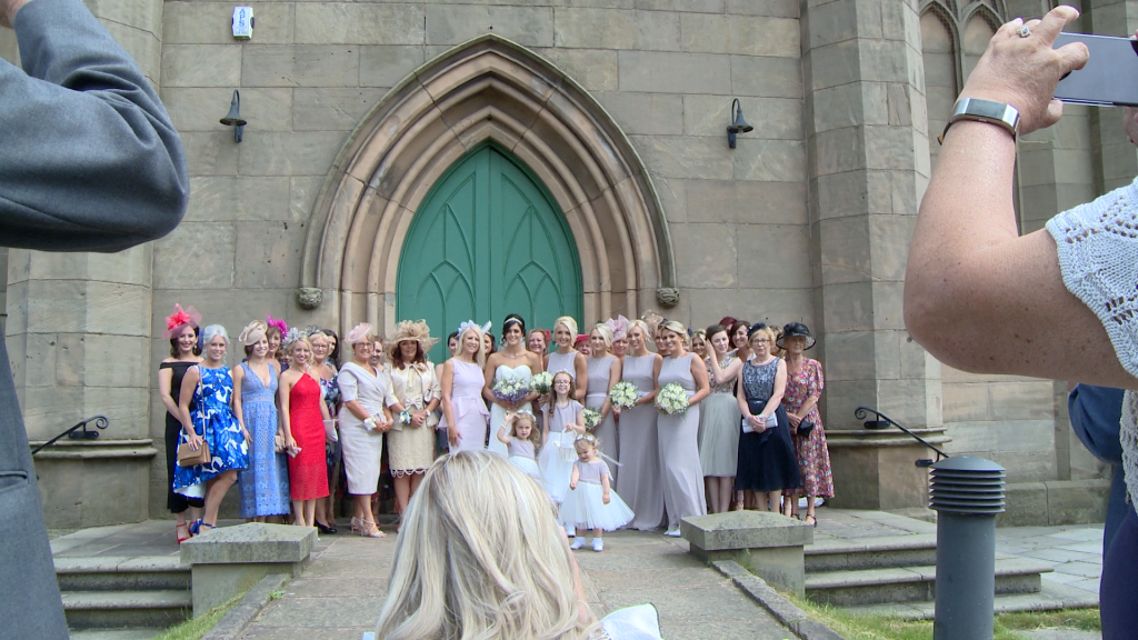 laura wade the wedding photographer ducks down to get a large group shot outside st johns church green doors as guests take photos on their smart phones