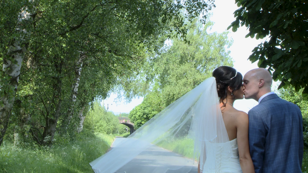 a bride and groom kiss on a country back lane between Burscough and Lathom in Lancashire. Her veil blows in the gentle breeze for the wedding photographer and videographer