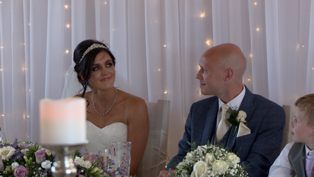 the bride tries not to cry during her dads father of bride speech at the blue mallard. They're sat in front of a white tulle backdrop with fairy lights and have purple and cream flowers for their colour scheme