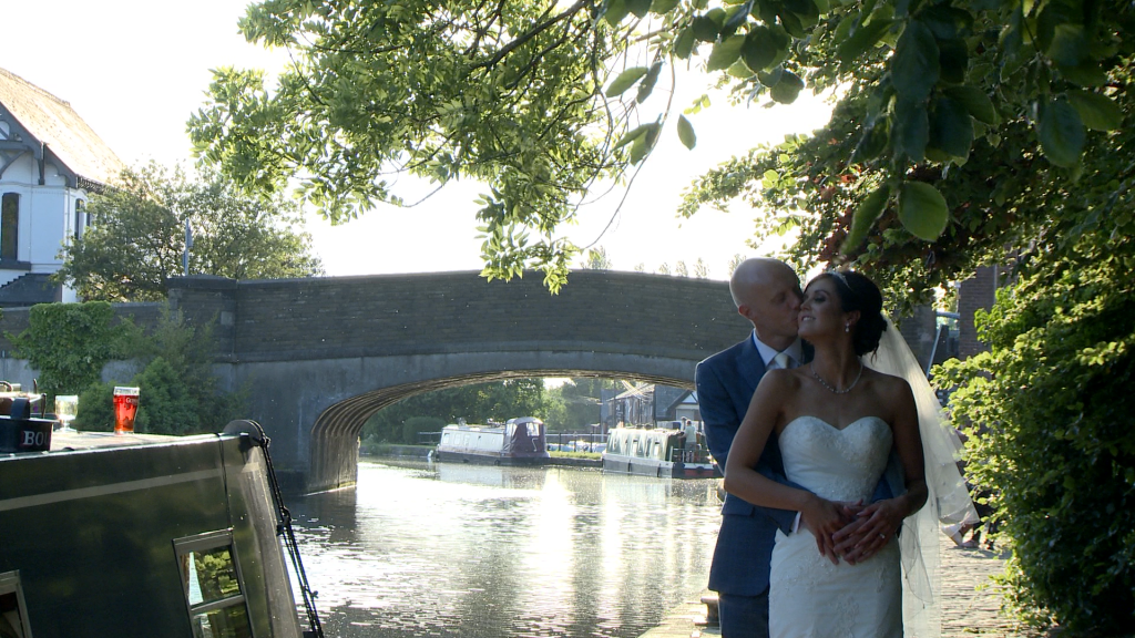 the bride and groom stand on the tow path by the canal under a shady tree with the canal bridge in the background