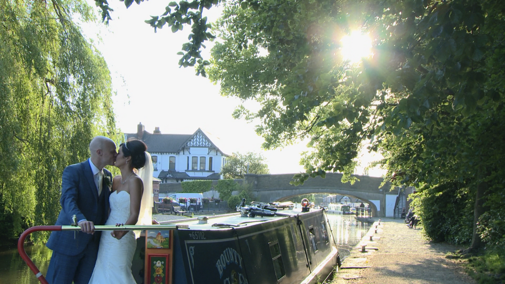 the bride and groom have a kiss standing on a canal boat outside Burscough wharf as the sun peeks through the trees for their wedding video