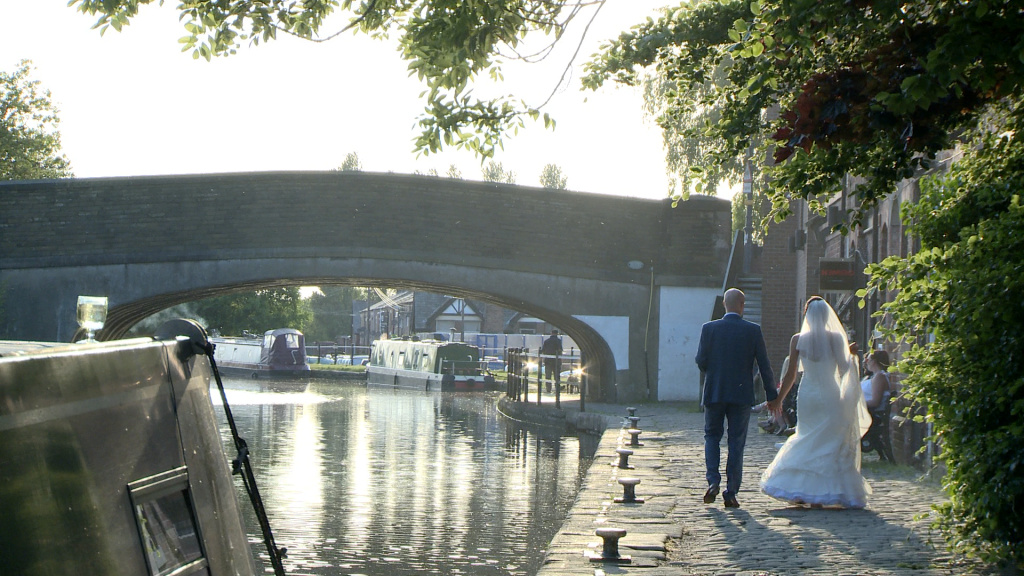 A video still from Lover Gets Sweeter wedding videography of the bride and groom walking back towards the Blue mallard down the tow path of the canal as the sun begins to set behind the trees in Burscough Lancashire