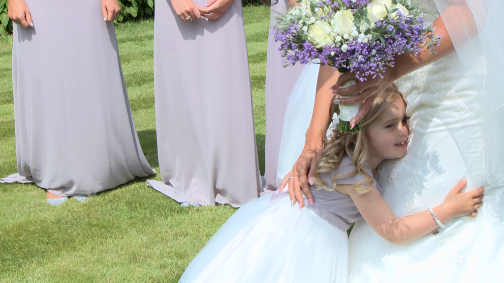 a flower girl wearing white and lilac with flowers in her hair gives her Mum in her wedding dress a big cuddle as the bridesmaids smile standing in her parents garden in Holmeswood whilst the sun shines during their summer wedding in Lancashire