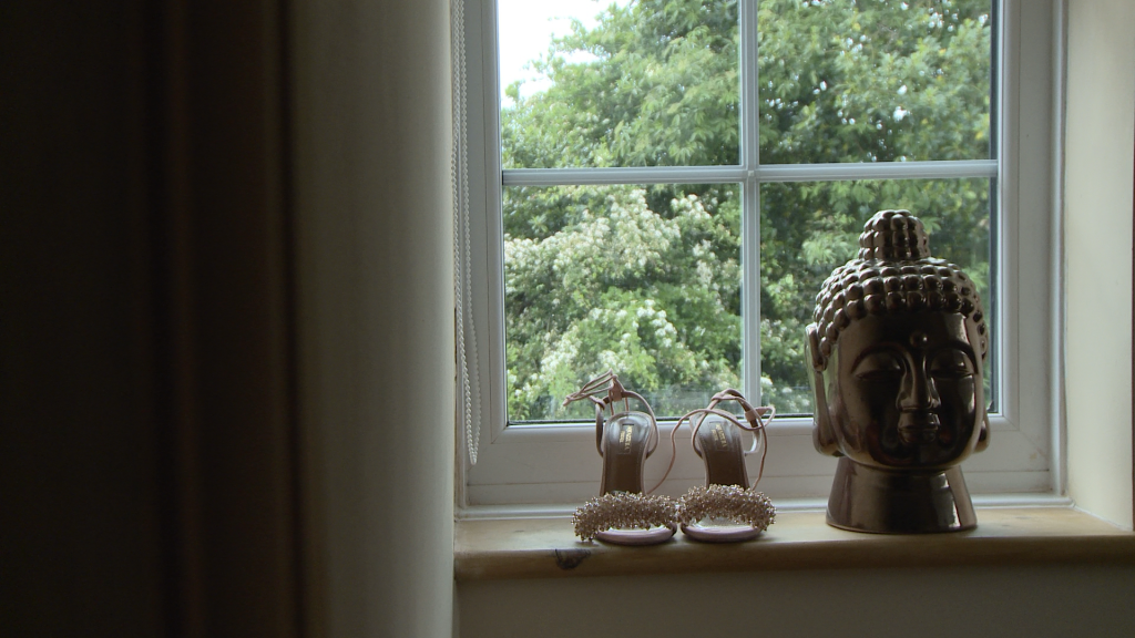 a pair of blush pink beaded bridal shoes sit on a window sill next to a bronzed Buddha head