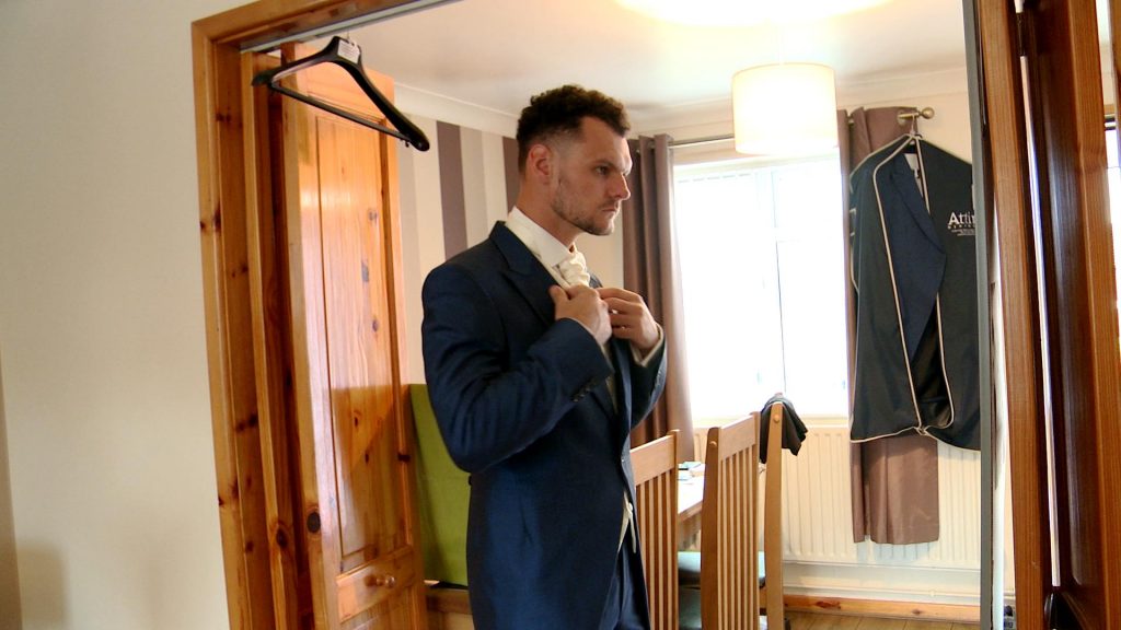 the groom looks in a mirror as he straightens his ivory tie and navy suit at home