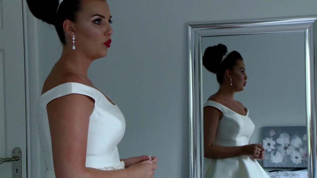 a classic old hollywood style bride takes a deep breath pouting her red lips with her reflection in the mirror behind her on the wedding video