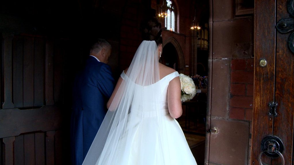 the bride links her dad's arm in the doorway of St Saviours church before walking down the aisle captured on her wedding video