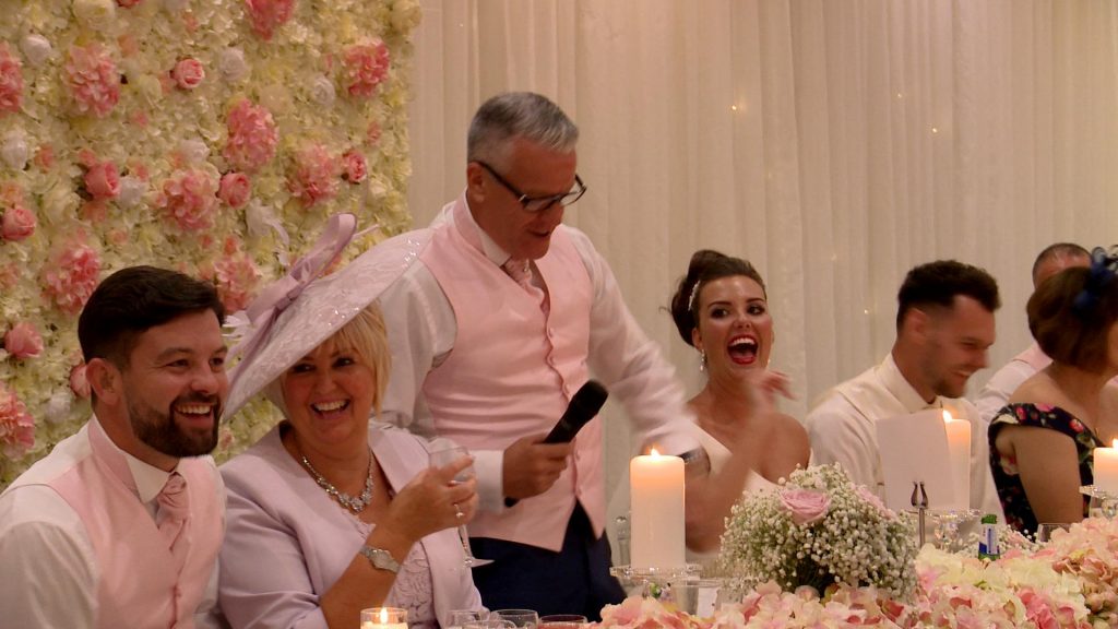 the father of the bride has everyone laughing during his wedding speech in front of a stunning pink and ivory flower wall at thornton hall