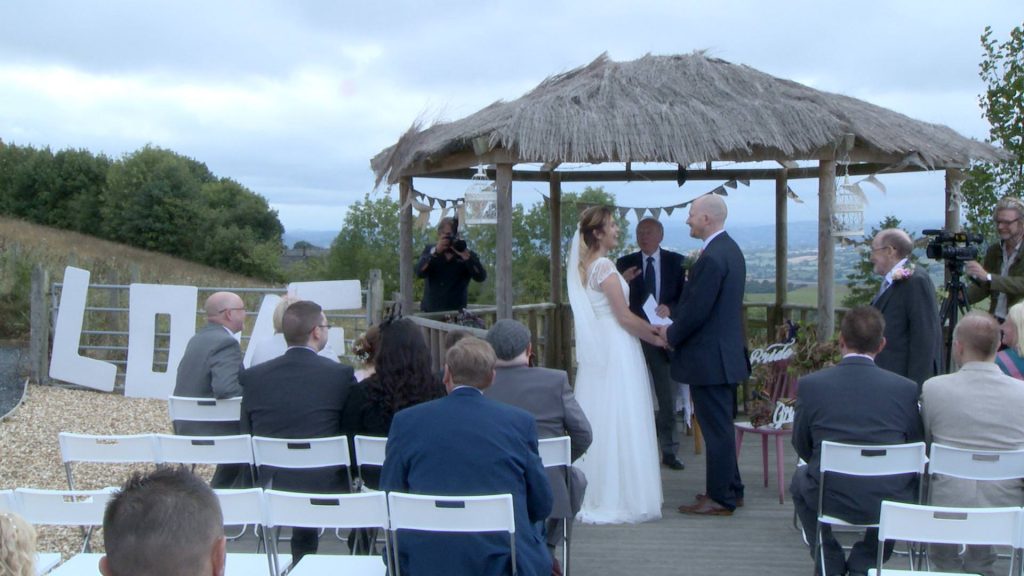 a video still of an outside ceremony at abel's harp in Shrewsbury with the bride and groom holding hands to say their wedding vows