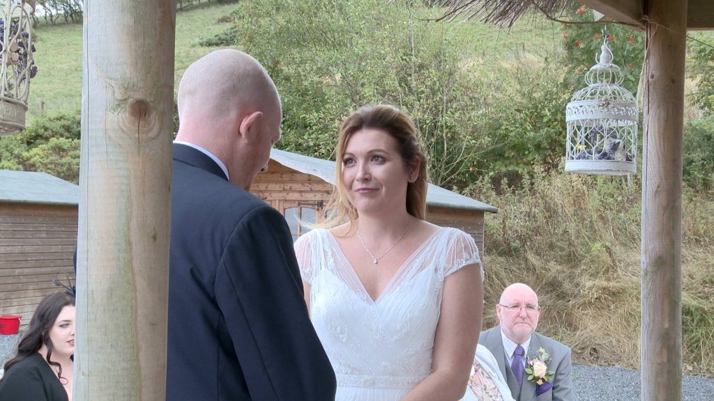 the bride looks lovingly at her husband for the wedding video during their outdoor ceremony