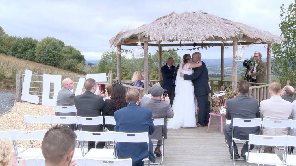 the bride and groom kiss as guests sat in the white sites during the outdoor ceremony at Abel's harp all clap and cheer for the wedding video