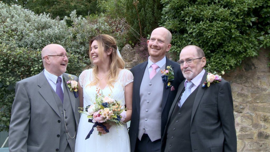 a bride smiles at her Dad during a group shot as the Groom looks at the camera with his dad