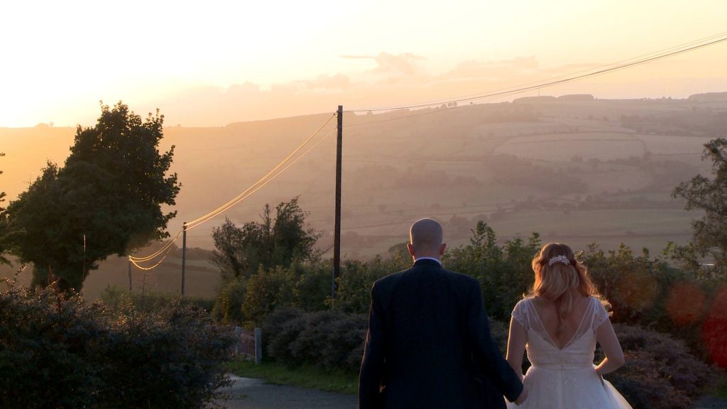 a stunning light peeks over the welsh hills as a bride and groom walk away from the videographer holding hands