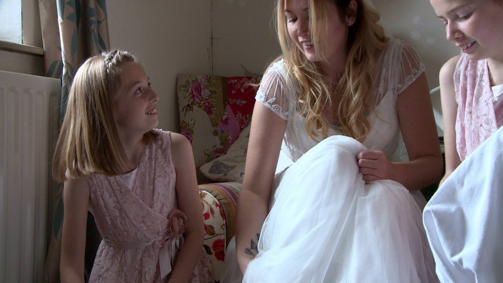 two flower girl daughters laugh with their mum as they try to help her get her white glitter irregular choice wedding shoes on for the videographer