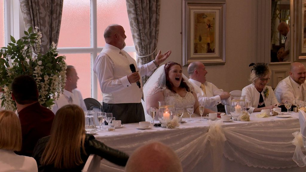 the father of the bride stands at the top table to do his wedding speech at Alma lodge and gets lots of reactions and laughter from the wedding guests and bride for their wedding video