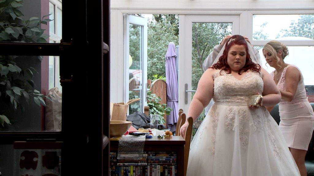 a bride with bright red hair stands holding a chair in her parents conservatory in Stockport as she has her wedding veil fitted before she gets married at Alma Lodge