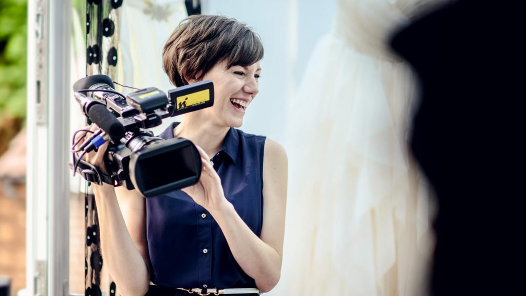 female videographer love gets sweeter relaxed and laughing with the bride and photographer Mick Cookson holding her Sony camera during filming on the morning of the brides wedding in Cheshire with her wedding dress hanging beautifully in the background