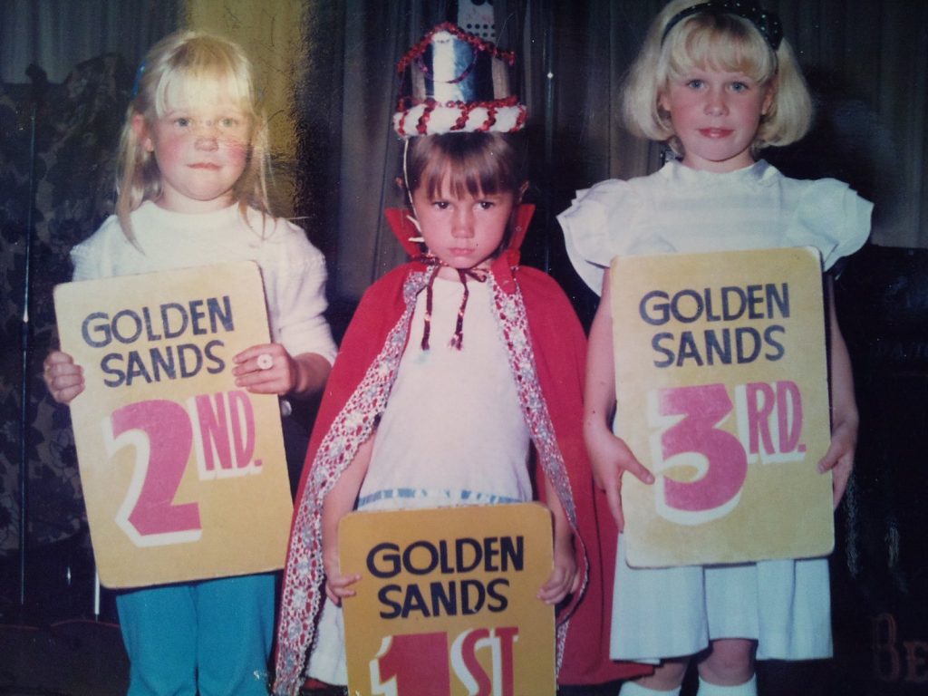 a photo of 3 little girls in a golden sands skegness beauty competition and the winner in the middle has a pout and red cloak on doesn't look happy about winning
